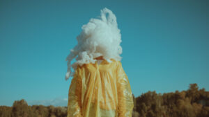 Henkilö keltaisessa takissa, pään kohdalla pumpulia symboloimassa pilveä.