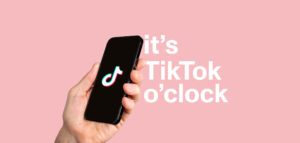 Hand holding a phone with TikTok logo on it and a text that says "It's TikTok o'clock" on a pink background.
